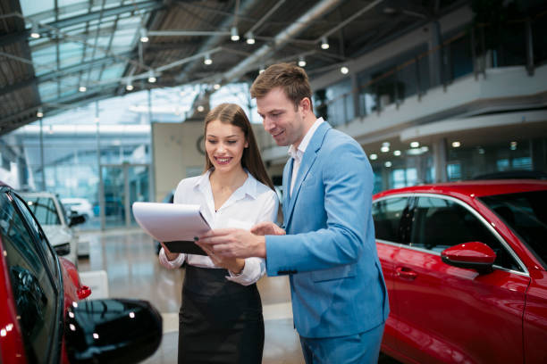 Smiling young saleswoman showing a car contract to male customer at new car showroom. Businessman looking at documents shown by female sales manager at automobile dealership.